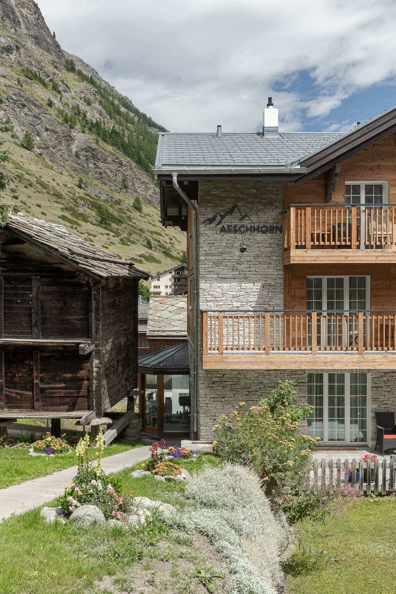 Seitliche Frontalansicht des Chalet Aeschhorn mit Naturstein- und Holzfassade, zentraler verglaster Eingang. Rechts traditionelle Holzhütte auf Steinsockeln, davor gepflasterter Weg und bunte Blumenbeete.