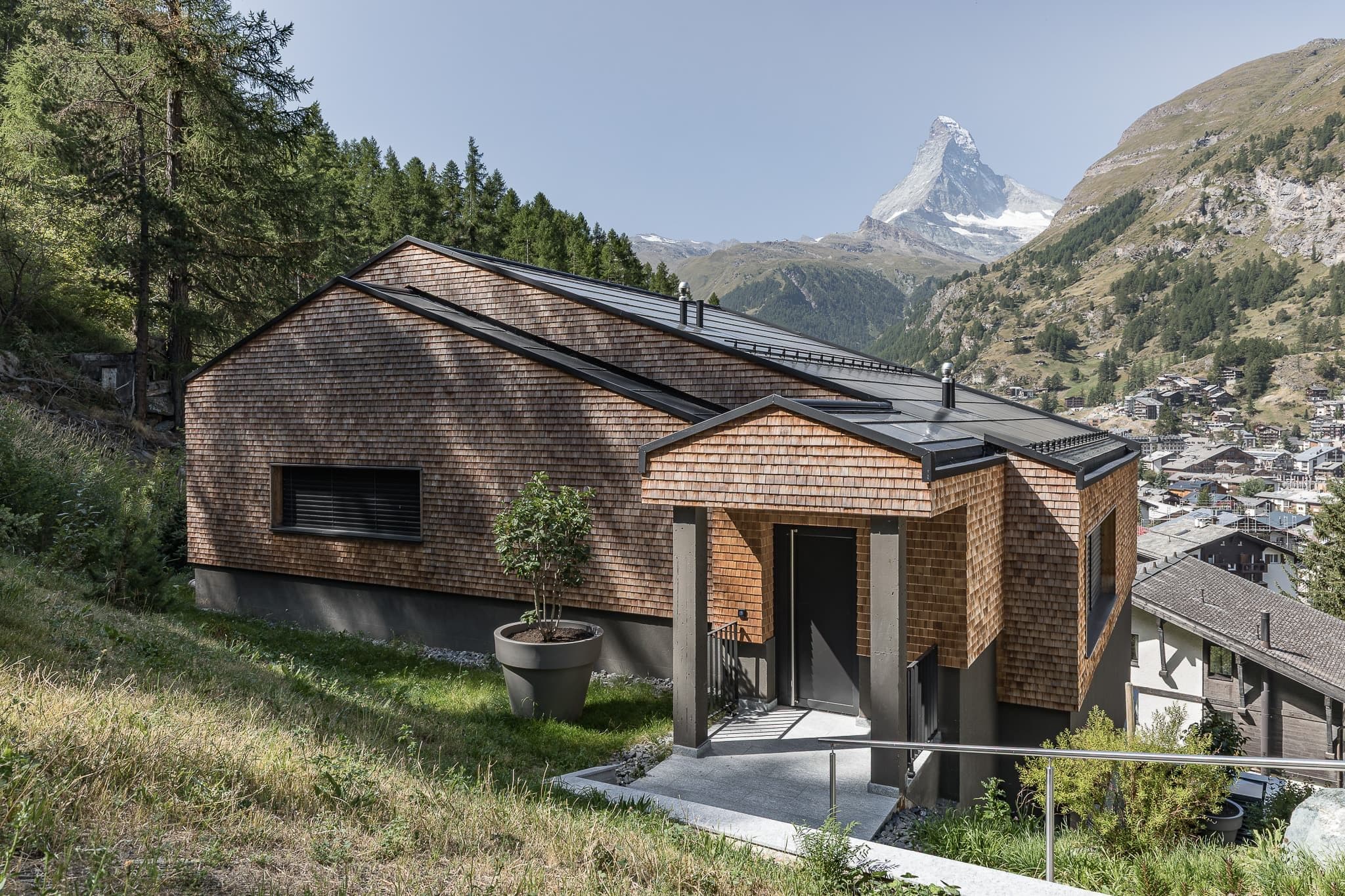 Aussenansicht des Chalets Pilou in Zermatt mit einer Fassade aus Holzschindeln und klaren, geometrischen Linien. Das Gebäude in moderner Formensprache liegt an einem Hang oberhalb von Zermatt und bietet einen direkten Blick auf das Matterhorn.